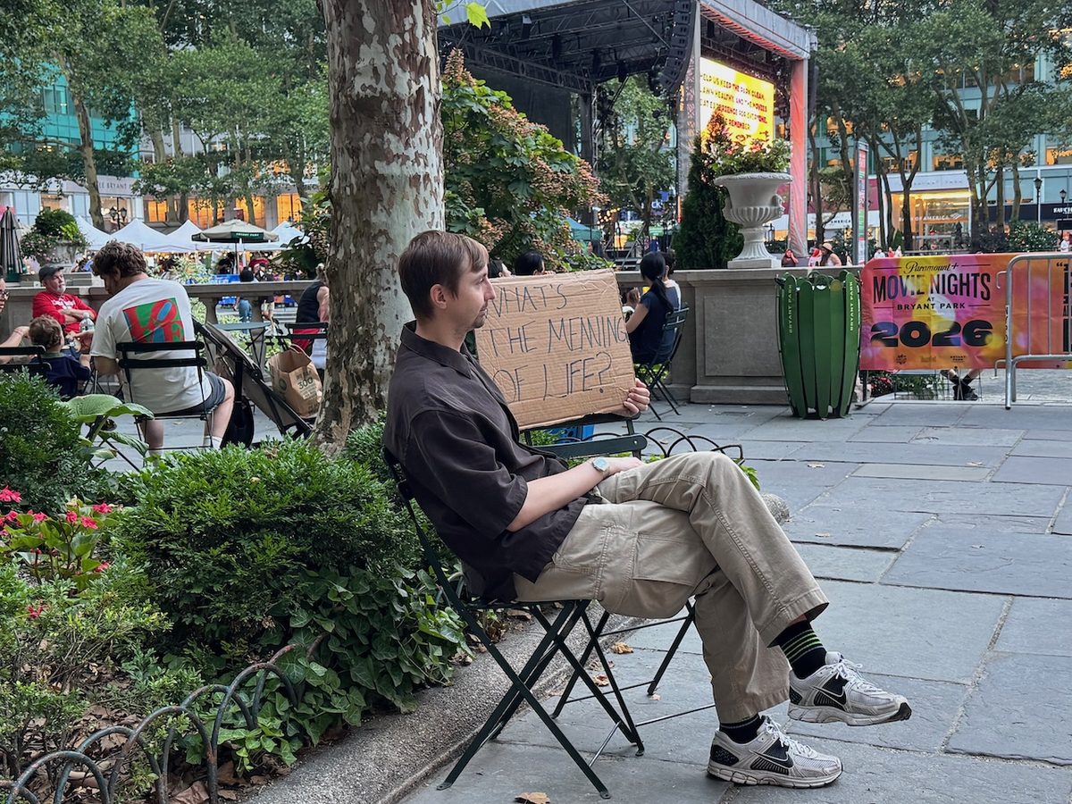 Alexey sitting in a park and holding a cardboard sign that reads “What’s the meaning of life?”